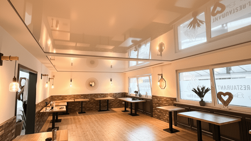 Moderner Restaurant-Gastraum in Menden mit einer spiegelnden weißen Hochglanz-Akustik Spanndecke, hellen Holztischen auf schwarzen Metallgestellen und einem Holzdielenboden. Die Decke reflektiert die warmen Edison-Glühbirnen-Pendelleuchten und die großen Fenster mit dem Schriftzug „Restaurant“ und Herz-Deko. Die Wände sind unten mit hellen Naturstein-Riemchen und oben mit weißem Putz gestaltet, verziert mit einem runden Sonnen-Akzentspiegel. Eine gemütliche, einladende Atmosphäre in Deutschland.